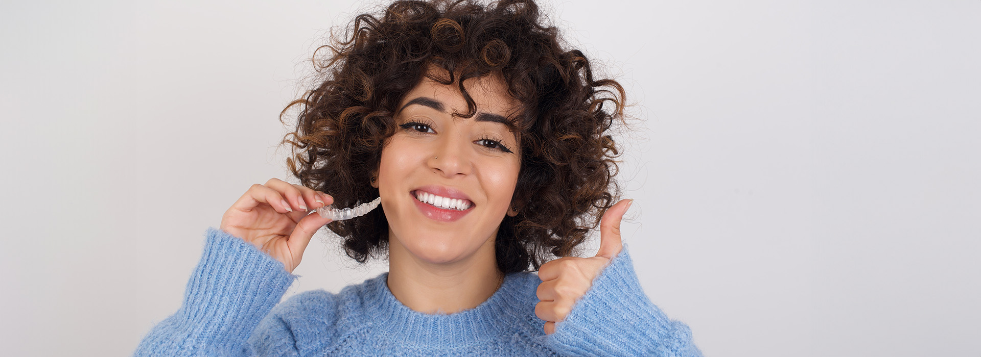 A woman with curly hair gives a thumbs-up gesture in front of a plain background.