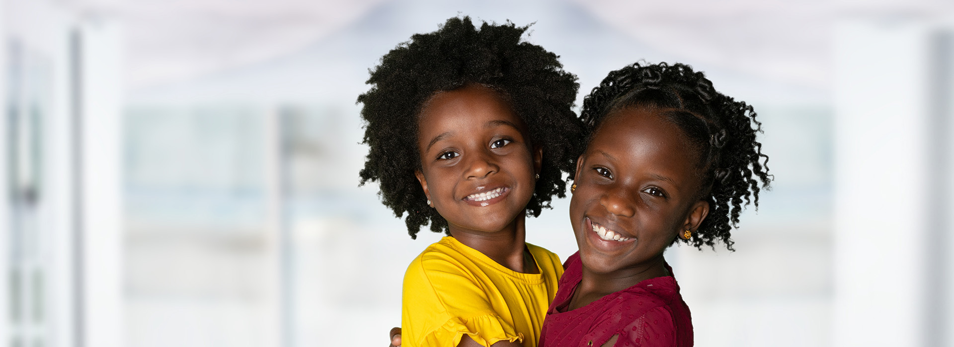 Two children smiling at the camera, one standing and one sitting, against a blurred background.