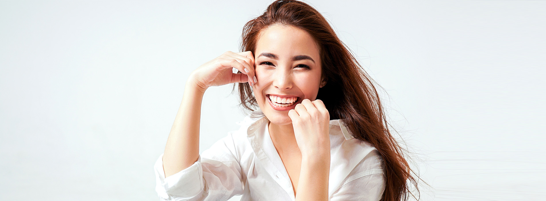 The image shows a smiling woman with long hair, holding her hands together near her face. She is wearing a light-colored top and appears to be in a studio setting with a blurred background that suggests she is the main subject of the photograph.