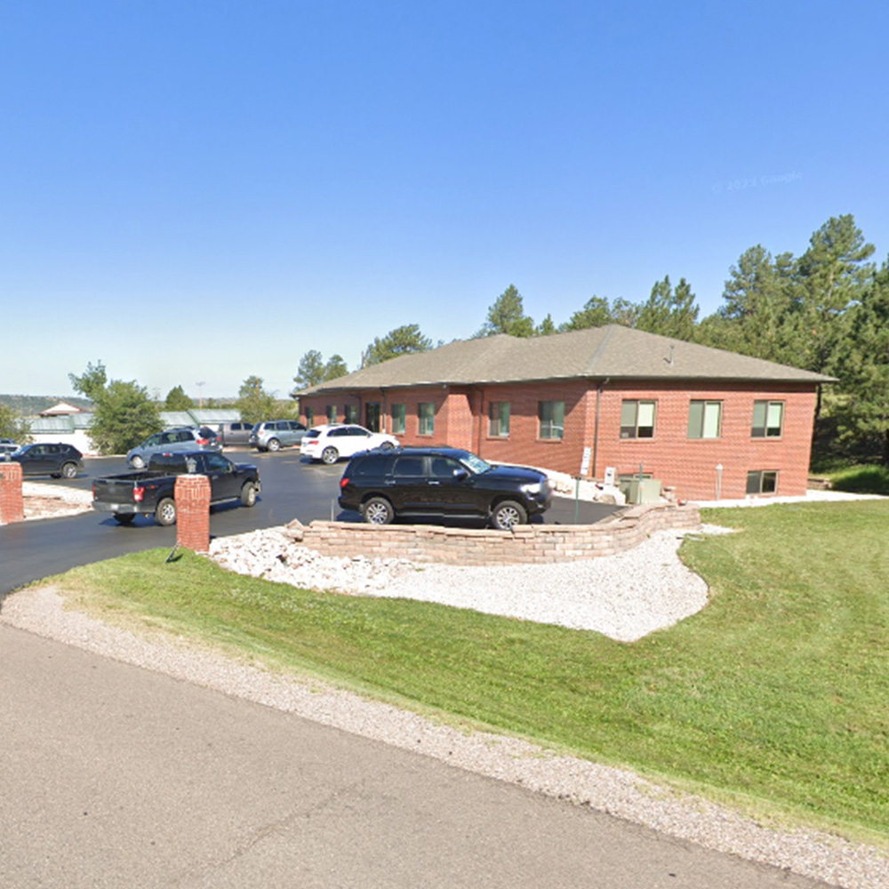 The image is of a large, red brick building with a parking lot, and there are multiple cars parked in front of the building.