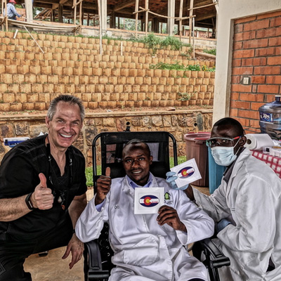 The image shows a man in a wheelchair holding up a sign with a logo on it, flanked by two men who appear to be medical professionals. They are smiling and giving thumbs-up signs, suggesting a positive or celebratory event.