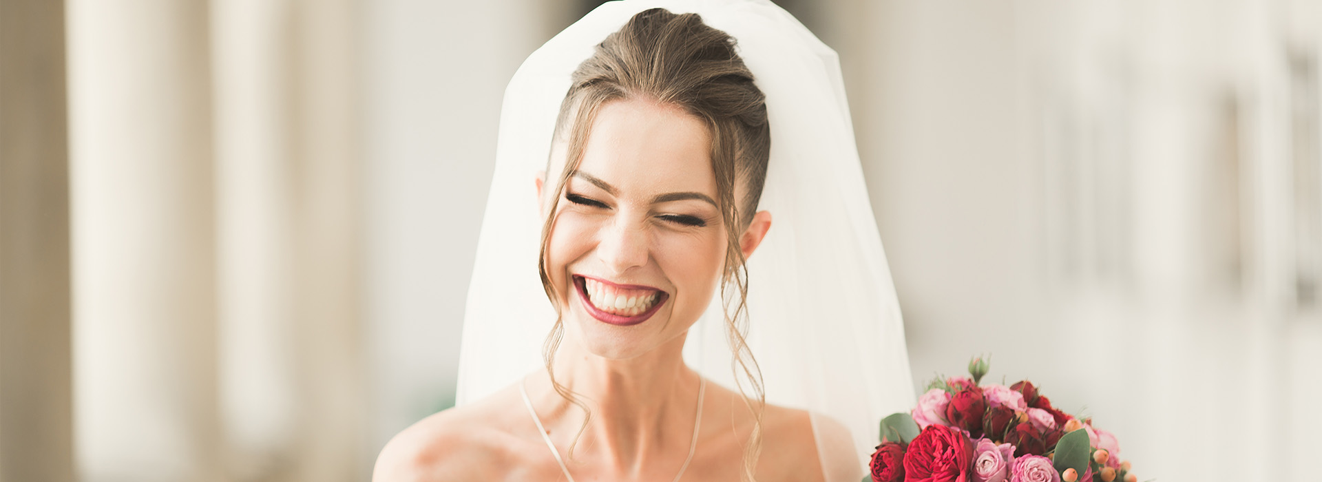 A bride in a white dress with a veil, holding a bouquet of flowers and laughing, standing in front of a building.